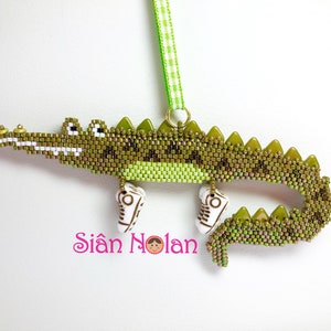 May include: A green beaded alligator wearing white sneakers, hanging from a green and white ribbon. The alligator is beaded with green, light green, and gold beads. The sneakers are white with black accents. The ribbon is tied at the top with a gold loop.