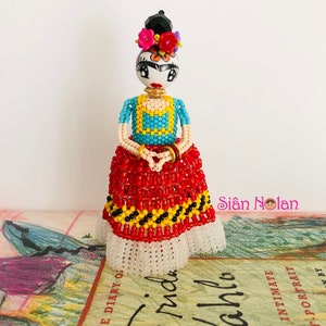 Puede incluir: Una mu&ntilde;eca de cuentas de Frida Kahlo, con una falda roja y negra, una blusa azul y amarilla y una corona de flores. La mu&ntilde;eca est&aacute; de pie sobre un libro titulado "El diario de Frida Kahlo".