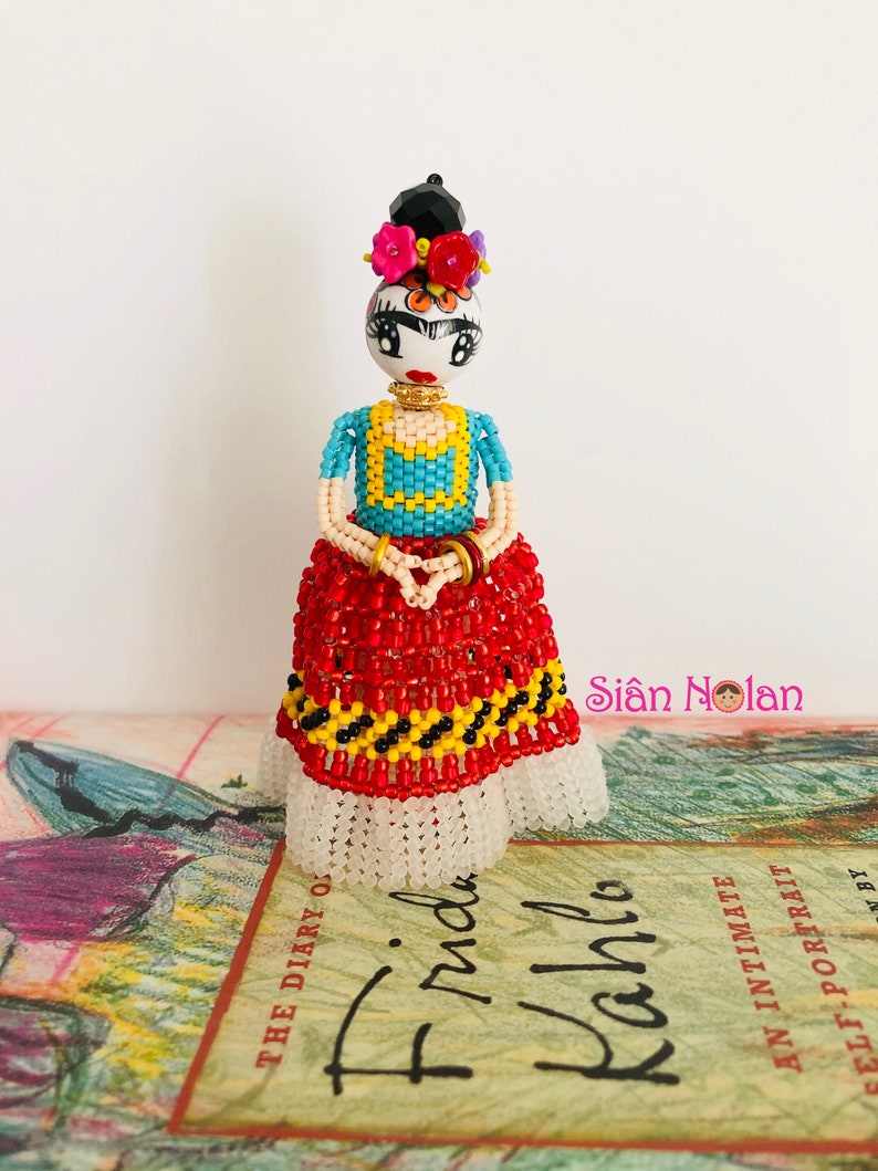 Puede incluir: Una mu&ntilde;eca de cuentas de Frida Kahlo, con una falda roja y negra, una blusa azul y amarilla y una corona de flores. La mu&ntilde;eca est&aacute; de pie sobre un libro titulado "El diario de Frida Kahlo".