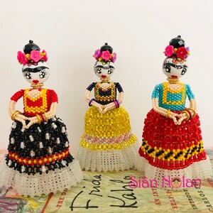 Puede incluir: Tres mu&ntilde;ecas de Frida Kahlo con cuentas, cabello negro, diademas florales de colores y vestidos de cuentas de colores. Las mu&ntilde;ecas est&aacute;n de pie sobre un libro con el t&iacute;tulo "Frida Kahlo" visible.