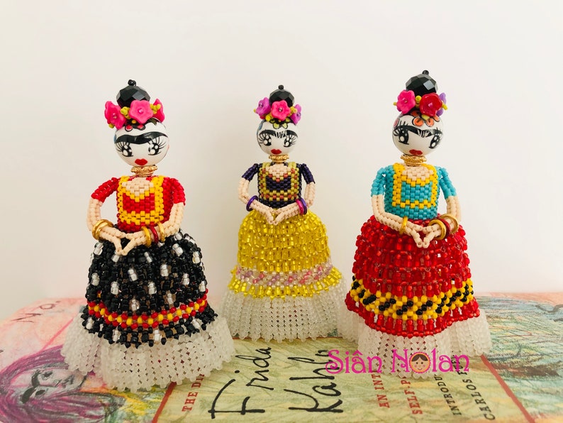 Puede incluir: Tres mu&ntilde;ecas de Frida Kahlo con cuentas, cabello negro, diademas florales de colores y vestidos de cuentas de colores. Las mu&ntilde;ecas est&aacute;n de pie sobre un libro con el t&iacute;tulo "Frida Kahlo" visible.