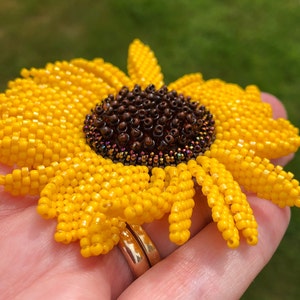 Könnte beinhalten: Eine handgefertigte Sonnenblumenbrosche aus Perlen. Die Brosche hat leuchtend gelbe Blütenblätter, die einen dunkelbraunen Mittelteil umgeben. Die Perlen sind sorgfältig angeordnet, um ein detailliertes Blumendesign zu erzeugen. Die Brosche wird in einer Hand vor einem verschwommenen grünen Hintergrund gehalten.