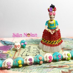 Peut inclure: Une poupée en perles en forme de crâne de sucre portant une robe rouge, jaune et noire avec une bordure en dentelle blanche. La poupée a des fleurs roses dans les cheveux et des yeux turquoise. La poupée est entourée d'une rangée de perles en forme de crâne de sucre blanches avec des fleurs turquoise et roses.
