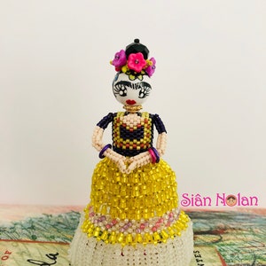 Puede incluir: Una mu&ntilde;eca de cuentas de Frida Kahlo con un vestido amarillo con ribete de cuentas blancas. La mu&ntilde;eca tiene un mo&ntilde;o negro con flores rosas y un collar de cuentas negras. La mu&ntilde;eca est&aacute; de pie sobre un libro con el texto "Frida" visible.