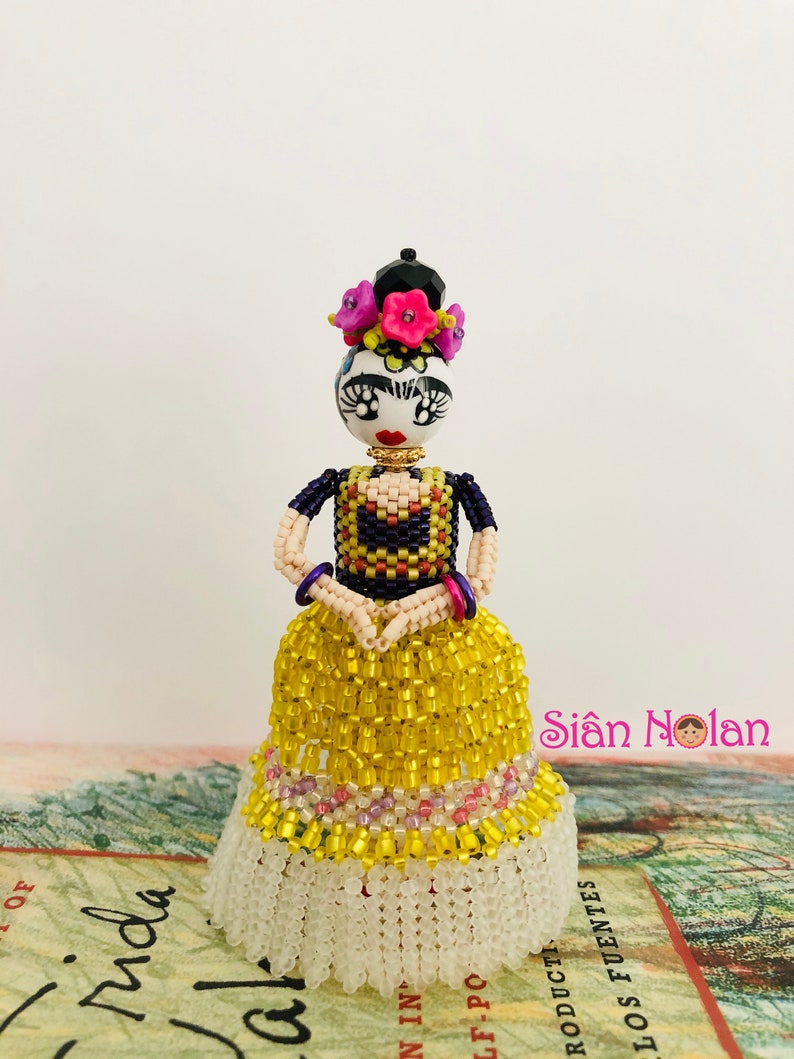 Puede incluir: Una mu&ntilde;eca de cuentas de Frida Kahlo con un vestido amarillo con ribete de cuentas blancas. La mu&ntilde;eca tiene un mo&ntilde;o negro con flores rosas y un collar de cuentas negras. La mu&ntilde;eca est&aacute; de pie sobre un libro con el texto "Frida" visible.