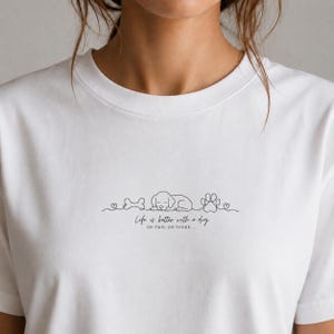 Puede incluir: Camiseta blanca con un dibujo lineal minimalista de un perro durmiendo, un hueso, una huella de pata y corazones. El texto "Life is better with a dog OR TWO, OR THREE..." está debajo. Estilo casual.