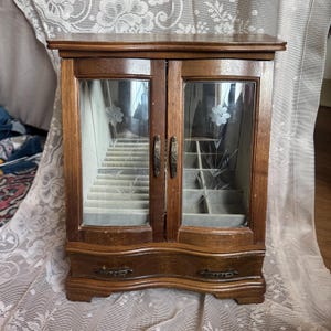 May include: A wooden jewelry box with two glass doors and a drawer. The doors have etched floral designs and open to reveal multiple compartments. The box is made of dark wood and has brass-colored handles and drawer pulls.