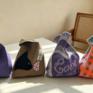 May include: Four small knit bags with knotted handles. The first is purple with a green duck graphic. The second is tan with a black cat and window design. The third is purple and white with the word Love. The fourth has a geometric orange and teal pattern.