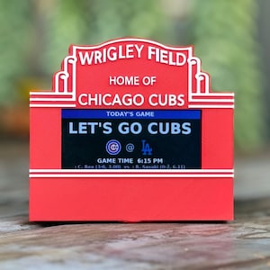 Peut inclure: Panneau rouge et blanc de Wrigley Field avec le texte "HOME OF CHICAGO CUBS" et "LET'S GO CUBS". Le panneau affiche l'heure du match et les équipes. Il s'agit d'un souvenir ou d'un objet de collection.