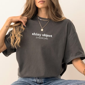 Puede incluir: Camiseta gris oscuro con las palabras "shiny object" sobre "syndrome" en letras blancas. La camiseta tiene mangas cortas y un corte holgado. La modelo lleva vaqueros azules y collares dorados.