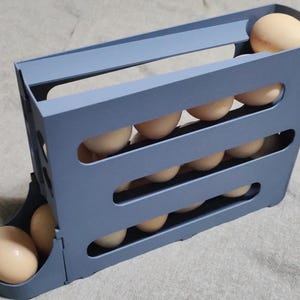 May include: A slate blue plastic egg dispenser featuring a tiered, gravity-fed design with multiple horizontal slots. The structure holds numerous light brown eggs, allowing them to roll down to the front tray for easy access and storage.