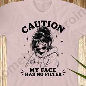 Könnte beinhalten: Ein hellrosa T-Shirt mit einer schwarzen Grafikillustration einer Person mit Dutt und Sonnenbrille. Der Text CAUTION ist über der Figur gewölbt, und der Text MY FACE HAS NO FILTER ist darunter gedruckt, umgeben von kleinen Sternsymbolen.