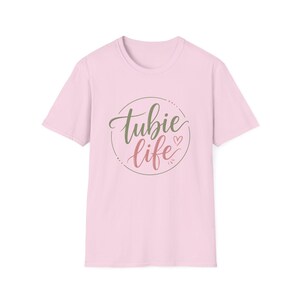 May include: A light pink short-sleeve t-shirt featuring a circular graphic on the chest. Inside the circle, the text 'tubie life' is written in a cursive script, with 'tubie' in olive green and 'life' in a muted rose color, alongside a small heart.