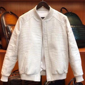 May include: Off-white bomber jacket with a textured, crocodile-like pattern. The jacket features a ribbed collar, cuffs, and hem, along with two front pockets. It is hanging on a wooden hanger against a backdrop of leather bags.