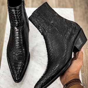 Könnte beinhalten: Zwei schwarze Leder-Ankle Boots mit Schlangenleder-Textur. Ein Stiefel steht aufrecht, der andere wird von einer Person gehalten. Die Stiefel haben spitze Zehen, seitliche Reißverschlüsse und kleine Blockabsätze. Die Stiefel stehen auf einer weißen Oberfläche.