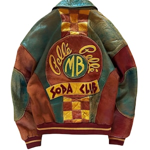 Puede incluir: Una chaqueta bomber de cuero multicolor con un gran parche circular en la espalda con el texto Pelle Pelle y MB, junto con un panel a cuadros amarillos y rojos. La chaqueta incluye puños acanalados de color burdeos y un dobladillo burdeos.