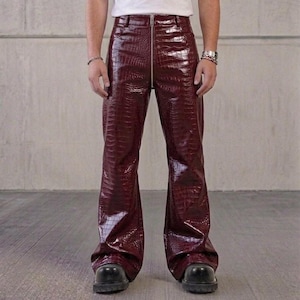 Op de afbeelding: Dieprode broek met een glanzende, in reliëf gemaakte krokodillenleertextuur. De broek heeft een hoge taille, een opvallende, zilverkleurige ritssluiting over de gehele lengte aan de voorzijde en een wijd uitlopend model.