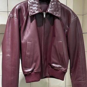Puede incluir: Una chaqueta de cuero color burdeos intenso con cuello de cuero trenzado, cierre central de cremallera metálica plateada, dos bolsillos verticales en la parte delantera y un dobladillo de punto acanalado en tono granate.