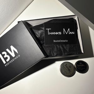May include: Black box with the text "13W BOOTSONTARIO" and "EVERY PAIR BEGINS A LEGACY". Inside the box is black tissue paper and a card saying "Thanks Man BootsOntario". Two silver shoe polish tins are also visible.