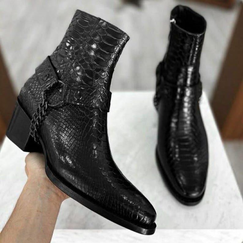 May include: Black leather ankle boots with a reptile-effect texture. The boots have a square toe, a small block heel, and a chain detail. One boot is held up, and the other is standing on a surface.