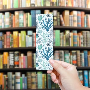 May include: A rectangular bookmark featuring a repeating pattern of blue and teal coral and seaweed designs on a white background. The bookmark is held in front of a blurred background of a bookshelf filled with books.