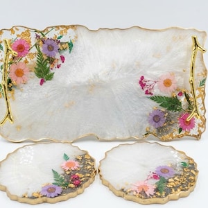 Puede incluir: Un juego de bandeja decorativa y dos posavasos de resina. El conjunto presenta una base blanca con flores secas incrustadas en rosa, morado y detalles dorados. La bandeja tiene asas doradas y un borde ondulado. Los posavasos tienen un diseño similar.