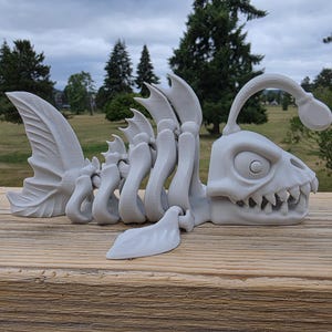 Könnte beinhalten: Eine hellgraue, 3D-gedruckte, bewegliche Anglerfisch-Skelettfigur. Das Wesen hat einen großen Kopf mit spitzen Zähnen, einen markanten Köder an der Stirn und einen segmentierten Körperbau, der eine flexible Bewegung des Objekts ermöglicht.