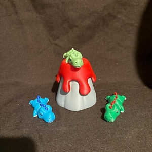 Puede incluir: Un modelo de volcán gris con lava roja goteando por los lados, con una pequeña figura de dinosaurio verde en la parte superior. Dos figuras de dinosaurio adicionales, una azul con púas azules y otra verde con púas rojas, están colocadas.