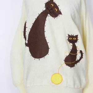 May include: A cream-colored knitted sweater with a brown cat and a smaller brown cat playing with a yellow ball of yarn.