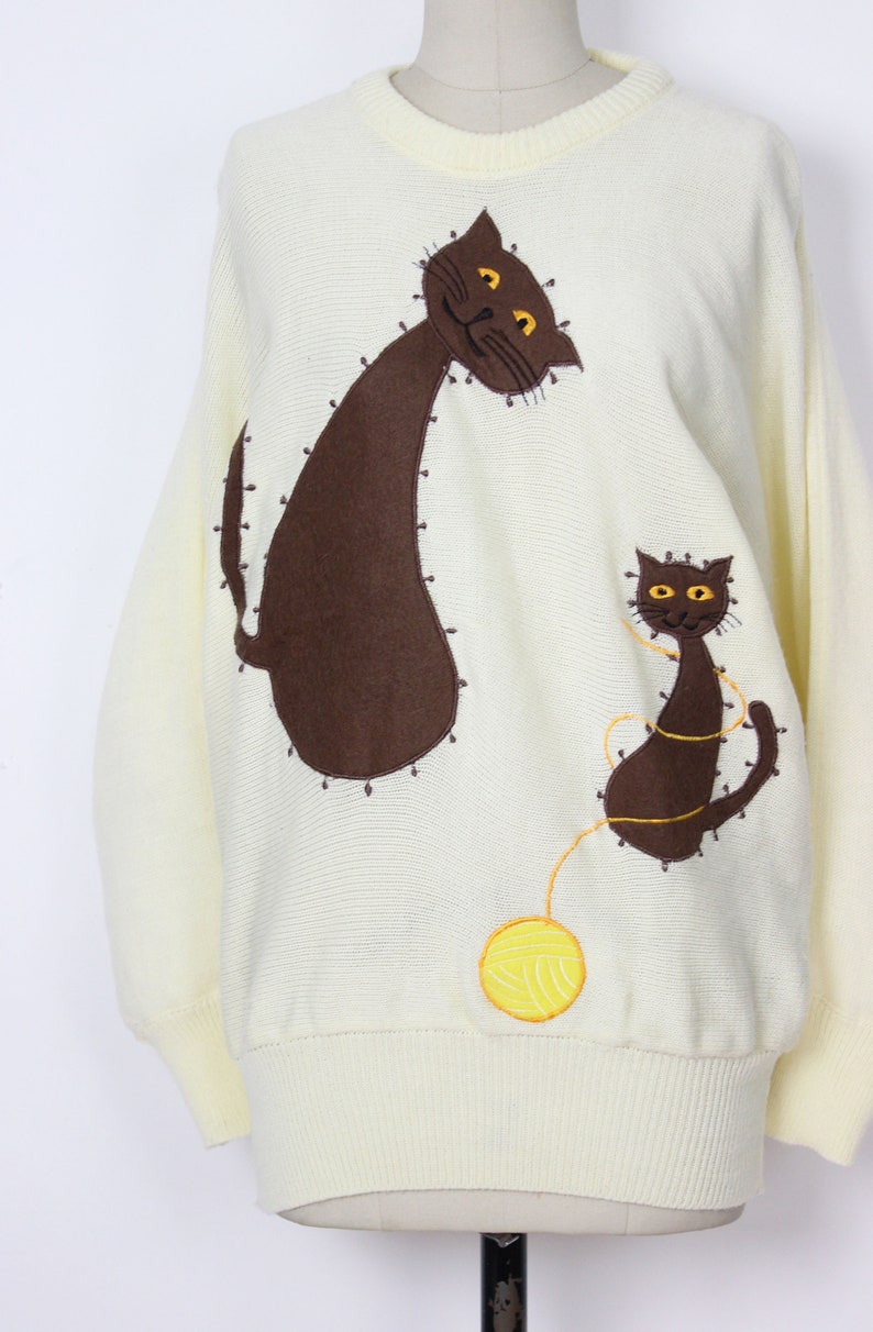 May include: A cream-colored knitted sweater with a brown cat and a smaller brown cat playing with a yellow ball of yarn.