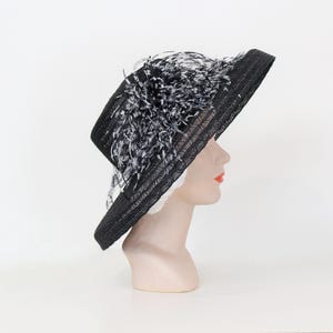 May include: A black sun hat with a wide brim and a decorative cluster of black and white feathers. The hat is displayed on a mannequin head, showcasing its shape and design. The hat's brim is approximately 4 inches wide.