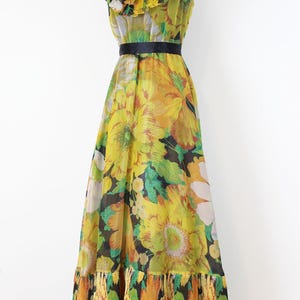Może przedstawiać: Wzorzysta sukienka maxi w stylu vintage na manekinie. Sukienka ma ż&oacute;łty, zielony i czarny wz&oacute;r kwiatowy, falbankowy dekolt, czarny pasek w talii i warstwową, plisowaną dolną krawędź. Sukienka jest bez rękaw&oacute;w i ma zwiewną sylwetkę.