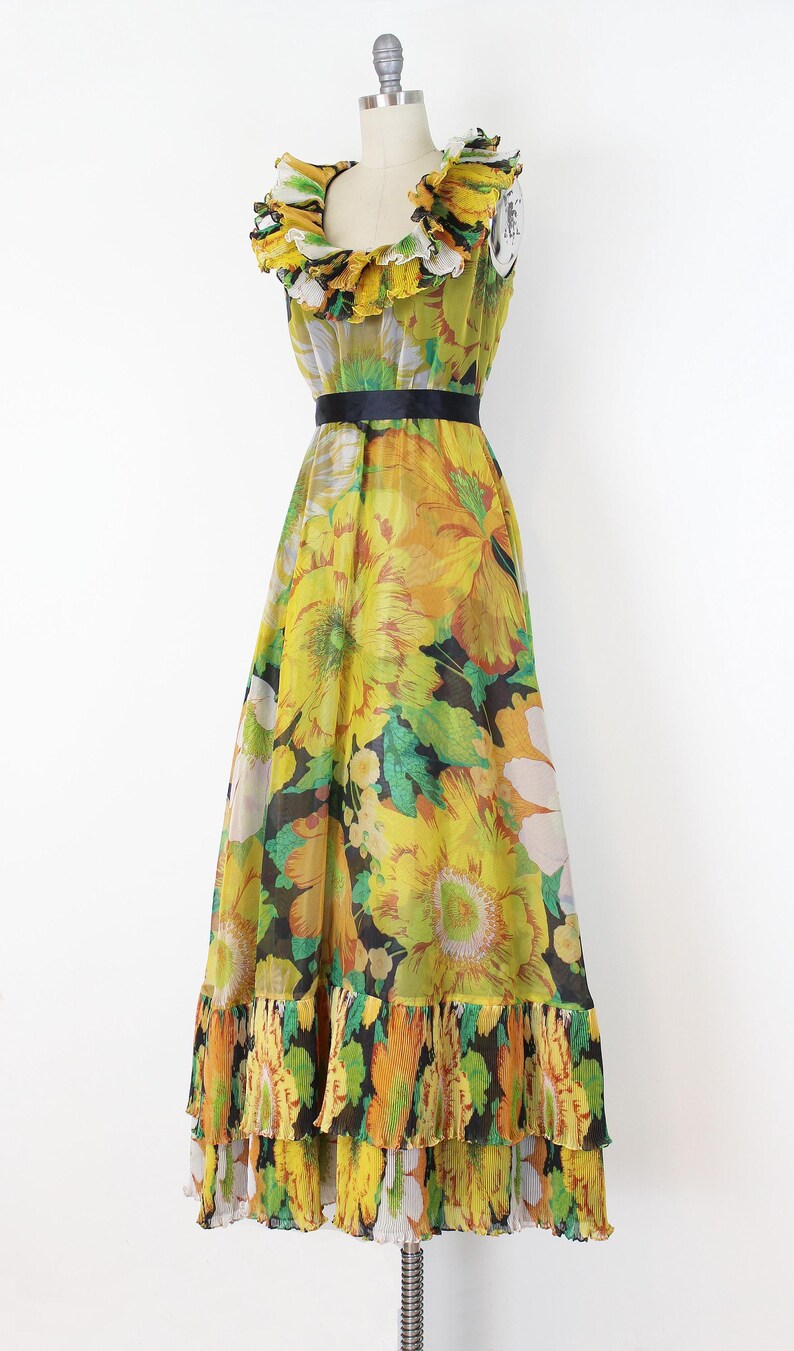 Może przedstawiać: Wzorzysta sukienka maxi w stylu vintage na manekinie. Sukienka ma ż&oacute;łty, zielony i czarny wz&oacute;r kwiatowy, falbankowy dekolt, czarny pasek w talii i warstwową, plisowaną dolną krawędź. Sukienka jest bez rękaw&oacute;w i ma zwiewną sylwetkę.