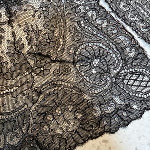 May include: Close-up of black lace fabric with intricate floral and paisley patterns. The delicate lace features a detailed design with a scalloped edge. The fabric is likely used for clothing or decorative purposes.