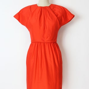 May include: A bright orange short-sleeved dress with a gathered neckline and a fitted bodice. The dress has a slightly flared skirt and a simple, elegant design.