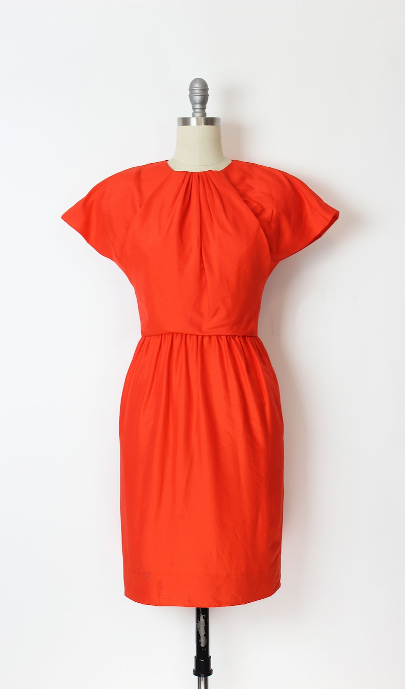 May include: A bright orange short-sleeved dress with a gathered neckline and a fitted bodice. The dress has a slightly flared skirt and a simple, elegant design.