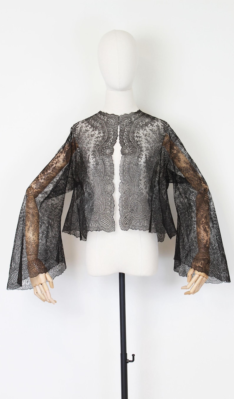 May include: A black lace jacket with wide, bell sleeves. The jacket is displayed on a mannequin against a white background. The intricate floral lace pattern is visible throughout the garment, creating a delicate and elegant look.