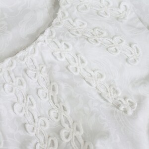 May include: Close-up of a white fabric with a delicate, repeating floral pattern. The edge of the fabric features an intricate, openwork trim with a repeating design. The overall aesthetic is soft and elegant, suggesting a vintage or handcrafted item.