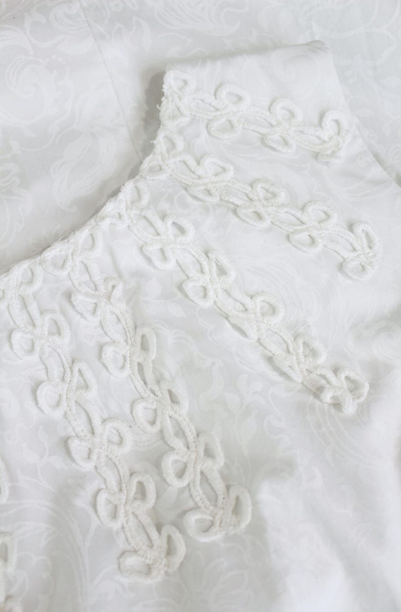 May include: Close-up of a white fabric with a delicate, repeating floral pattern. The edge of the fabric features an intricate, openwork trim with a repeating design. The overall aesthetic is soft and elegant, suggesting a vintage or handcrafted item.