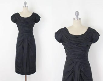 May include: A black dress with a scoop neckline and short sleeves. The bodice features gathered fabric, and the skirt has a draped design. The dress is displayed on a mannequin against a white background. A classic vintage style.
