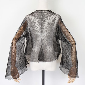 May include: Black lace top with long, flared sleeves. The sheer fabric reveals intricate floral patterns. The top is displayed on a mannequin against a white background, showcasing its delicate design and texture.