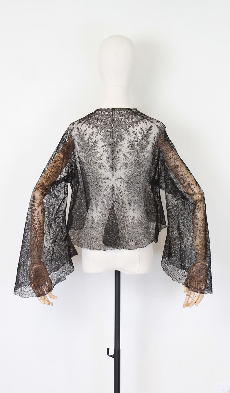 May include: Black lace top with long, flared sleeves. The sheer fabric reveals intricate floral patterns. The top is displayed on a mannequin against a white background, showcasing its delicate design and texture.