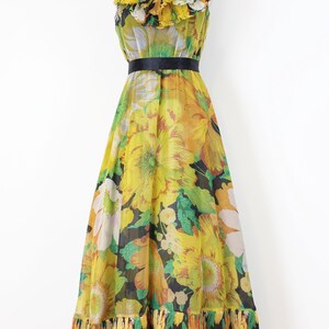 Może przedstawiać: Wzorzysta sukienka maxi w stylu vintage na manekinie. Sukienka ma ż&oacute;łty, pomarańczowy i zielony kwiatowy wz&oacute;r. Ma falbankowy dekolt, czarny pasek i warstwową, falbankową dolną krawędź. Sukienka jest w rozmiarze M.