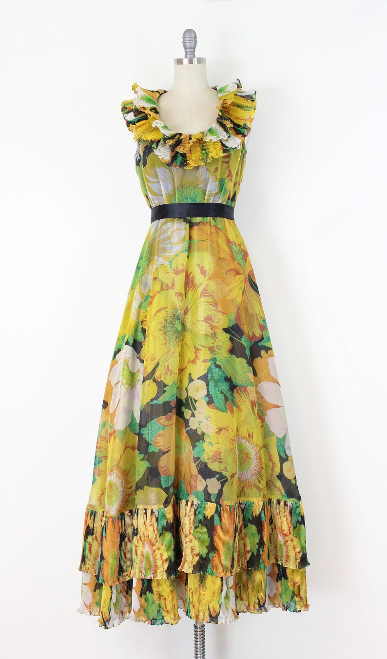 Może przedstawiać: Wzorzysta sukienka maxi w stylu vintage na manekinie. Sukienka ma ż&oacute;łty, pomarańczowy i zielony kwiatowy wz&oacute;r. Ma falbankowy dekolt, czarny pasek i warstwową, falbankową dolną krawędź. Sukienka jest w rozmiarze M.