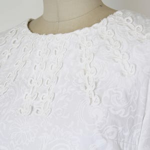 May include: White sleeveless dress with intricate floral and scrollwork embroidery around the neckline and bodice. The dress is made of a textured fabric, and the design is visible throughout the garment.