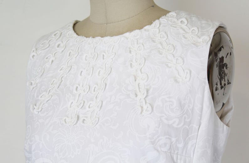 May include: White sleeveless dress with intricate floral and scrollwork embroidery around the neckline and bodice. The dress is made of a textured fabric, and the design is visible throughout the garment.