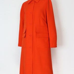 May include: A long, bright orange coat with a button-up front and two large pockets. The coat is on a mannequin.