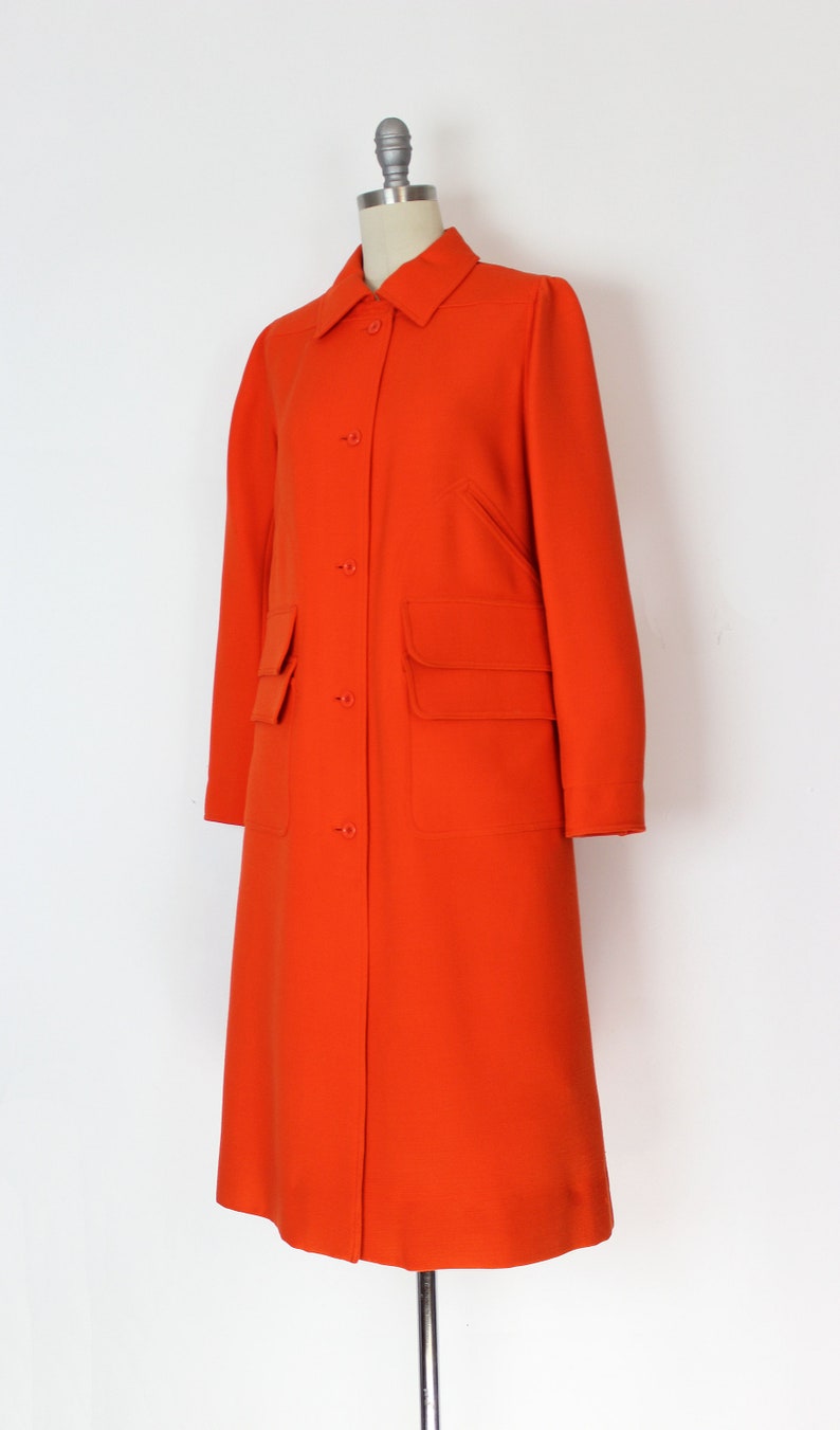 May include: A long, bright orange coat with a button-up front and two large pockets. The coat is on a mannequin.
