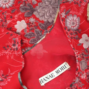 May include: Red sheer fabric with a floral print in shades of gray and white. The fabric has a ruffled edge and a label that reads "HANAE MORI".