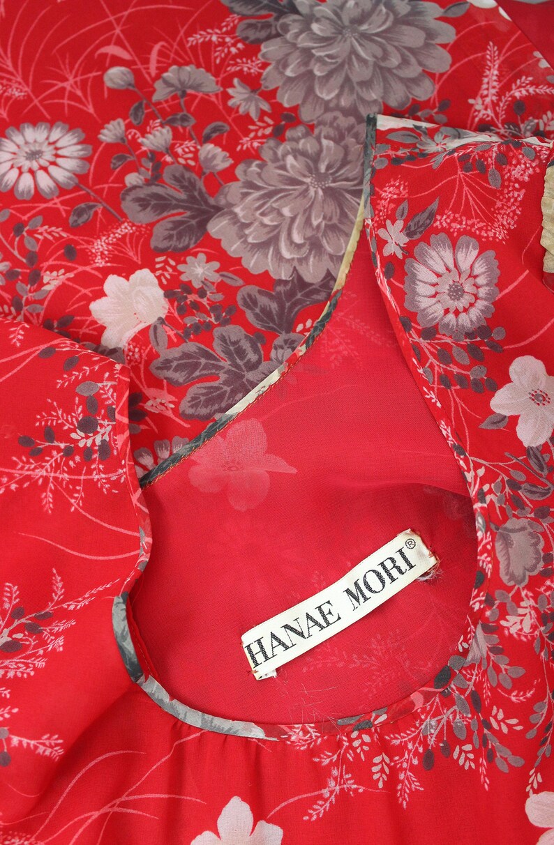 May include: Red sheer fabric with a floral print in shades of gray and white. The fabric has a ruffled edge and a label that reads "HANAE MORI".